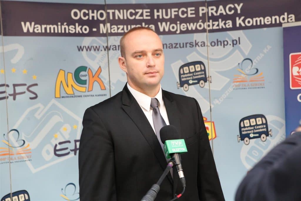 Mężczyzna w marynarce przemawia na tle baneru z nazwą targów w Centrum Konferencyjno-Szkoleniowym Uniwersytetu Warmińsko-Mazurskiego.
