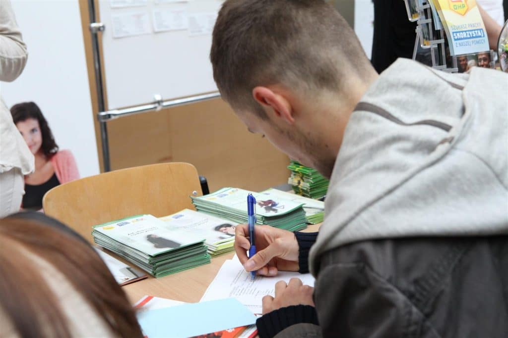 Młody mężczyzna zapisuje się na targach pracy, na stole wiele ulotek i materiałów promocyjnych.
