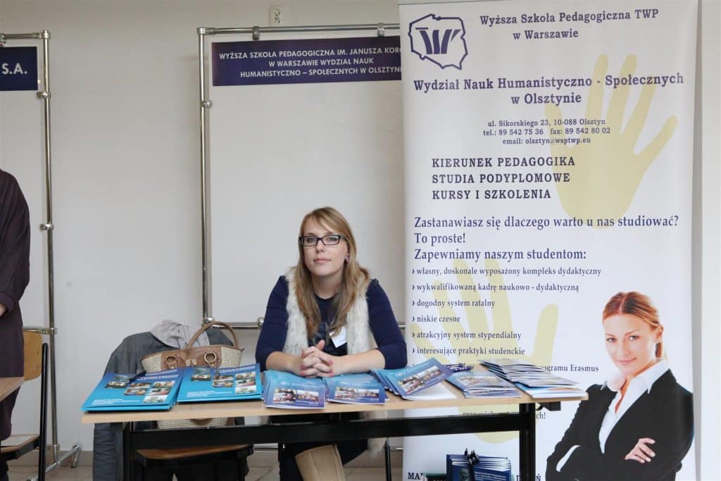 Stoi kobieta przy stoisku informacyjnym na targach pracy, za nią baner promujący kierunek pedagogika.