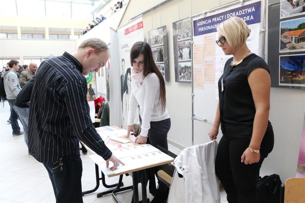Uczestnicy zwiedzają stoisko podczas targów pracy w Kortowie, przy stołach z materiałami i fotografiami.