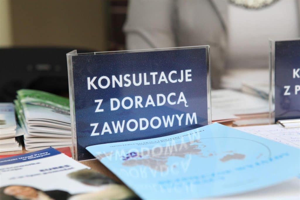 Stolik z materiałami informacyjnymi i oznaczeniem 'Konsultacje z doradcą zawodowym' przy stoisku na targach pracy.