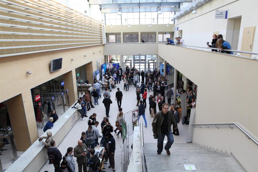 Widok na halę centrum konferencyjnego Uniwersytetu Warmińsko-Mazurskiego w trakcie targów pracy, ludzie rozmawiają i korzystają z ofert wystawców.