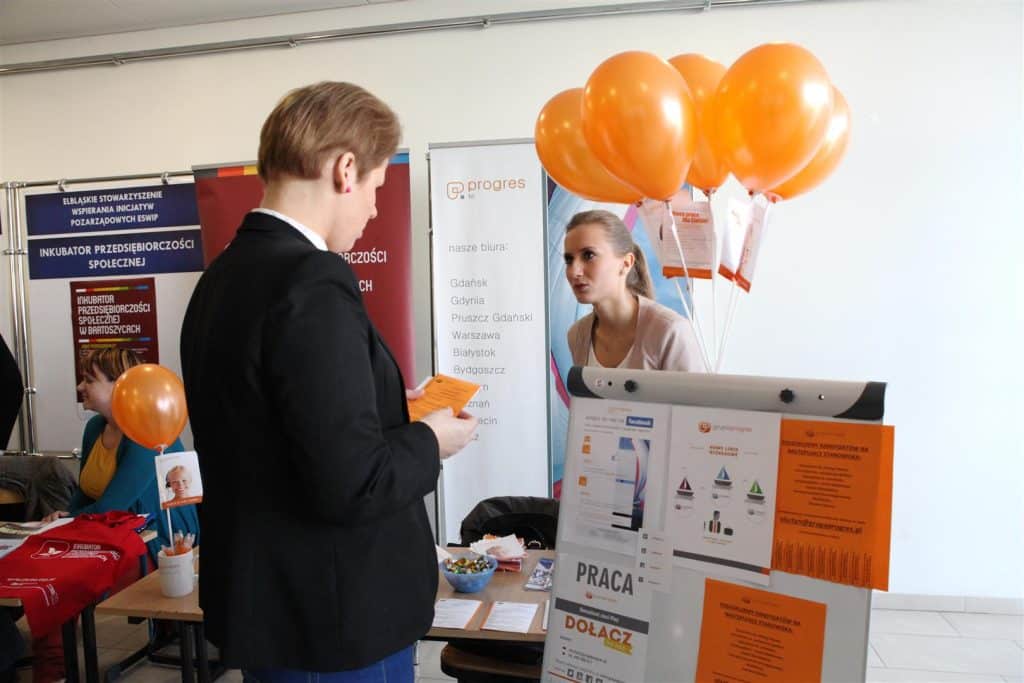 Młoda kobieta rozmawia z przedstawicielką stoisku podczas targów pracy w Kortowie, widoczne balony i materiały promocyjne.