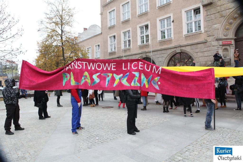 Uczniowie liceum plastycznego trzymają transparent z napisem 'Państwowe Liceum Plastyczna' na placu przy ratuszu w Olsztynie.