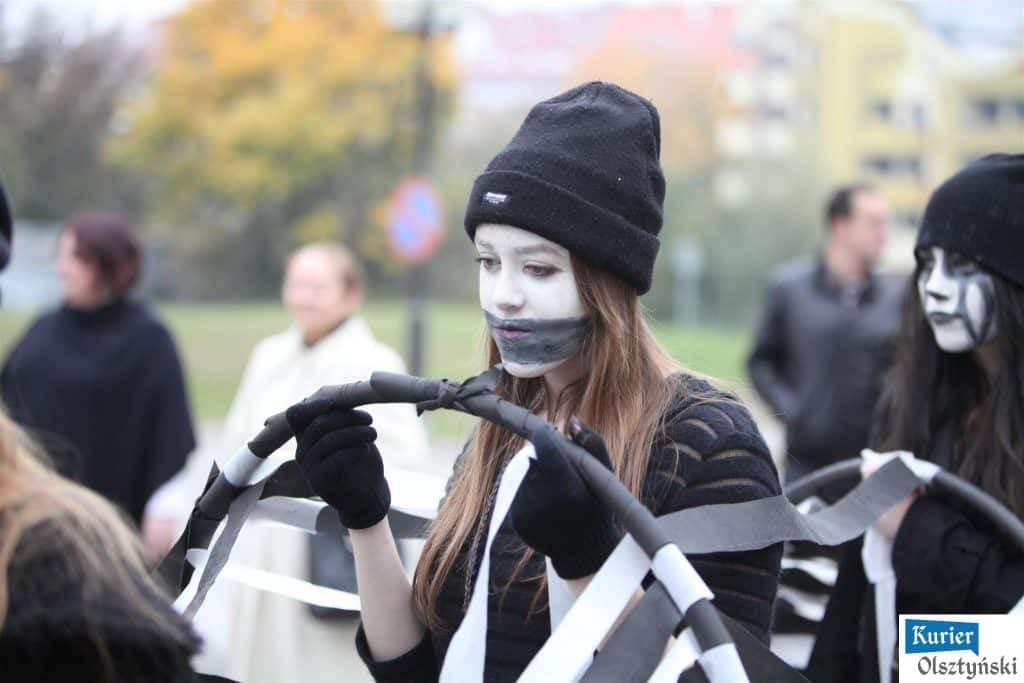 Uczennica z makijażem i ubraniem w czarno-białej tonacji trzyma taśmę, wokół niej inni w podobnym stylu.