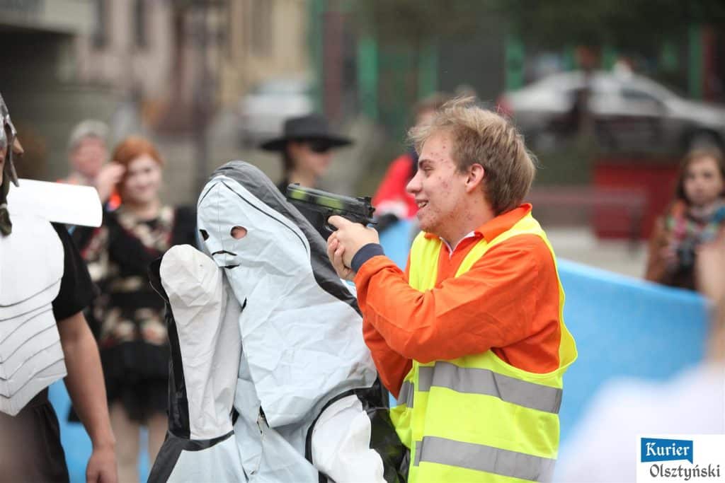 Młody uczestnik festiwalu celuje w stylizowaną postać z maską podczas wydarzenia plastycznego na placu.