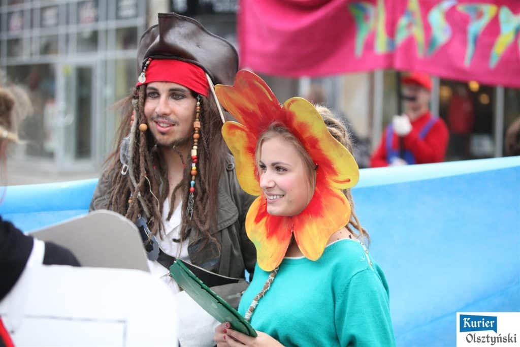 Uczestnicy festiwalu w kolorowych kostiumach na ulicy, jedna z nich z nakładką w kształcie kwiatu na głowie.
