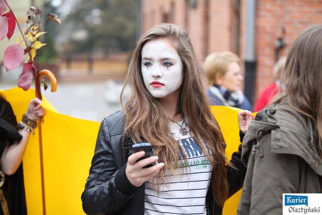 Uczennica plastycznego liceum z białym makijażem i ciemną kreską łez na twarzy, trzymająca telefon, podczas obchodów 25-lecia szkoły w Olsztynie.