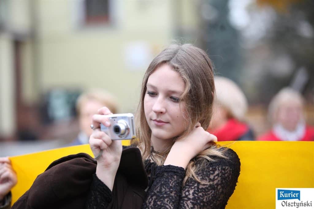 Uczennica w czerni fotografuje na ulicy, za nią rozmyte tłumy i budynki.