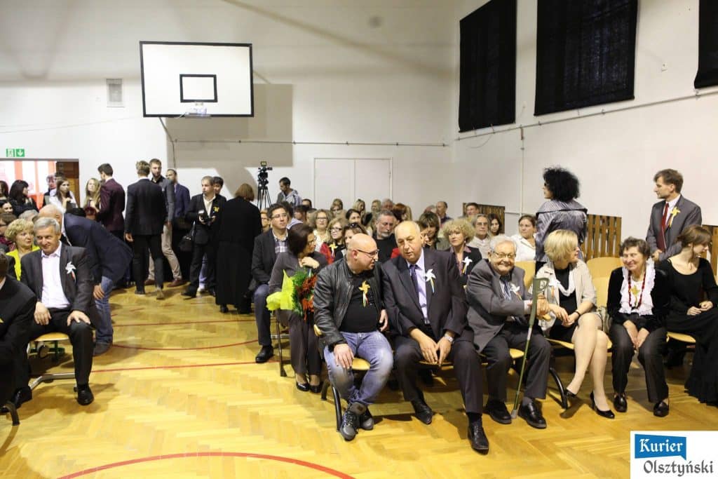 Uczestnicy uroczystości 25-lecia szkoły plastycznej siedzą i stoją w sali, przygotowują się do wydarzenia, w tle scena i kamera.
