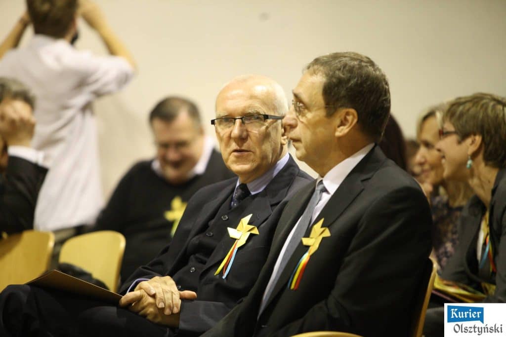 Uczestnicy uroczystości 25-lecia szkoły plastycznej, siedzą w sali, na pierwszy planie dwa mężczyźni w garniturach z ozdobami na piersiach.
