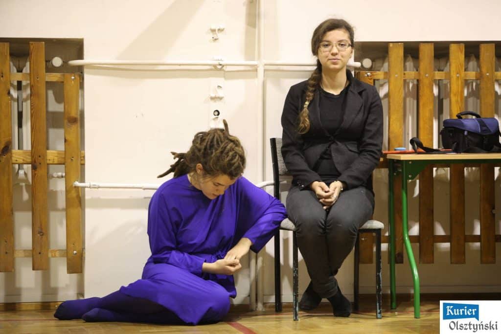 Dwie młode kobiety w trakcie występu na scenie, jedna siedzi na krześle, druga przechyla się do przodu, w tle drewniane elementy sceny.