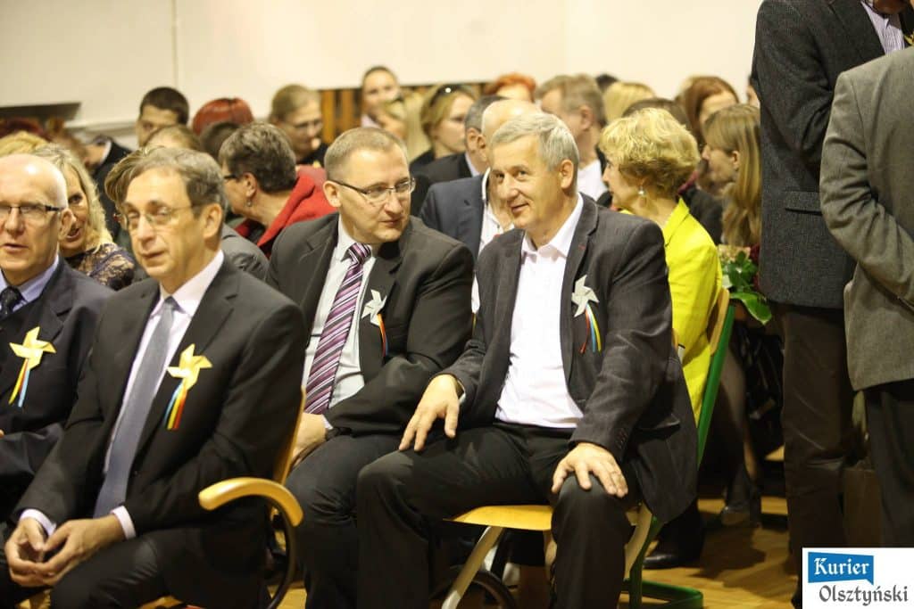 Uczestnicy uroczystości 25-lecia szkoły plastycznej, siedzą na sali, w tym mężczyźni w garniturach z dekoracyjnymi kokardami.