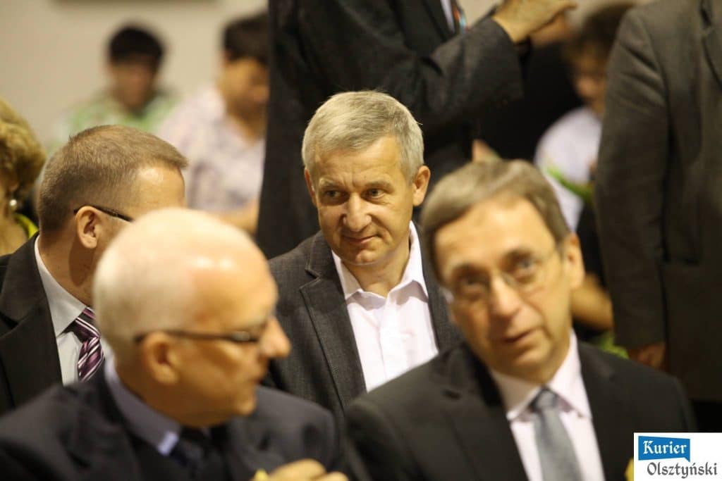 Grupa dorosłych mężczyzn siedzi na sali podczas uroczystości, w tle widoczni inni uczestnicy.