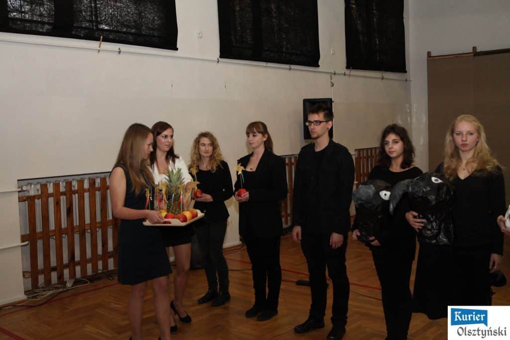 Grupa uczniów w sali szkolnej, jeden z nich trzyma kosz z symbolami owoców i dekoracjami, inni z napięciem patrzą.