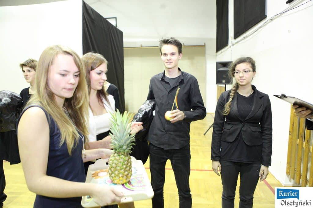 Uczniowie i nauczyciele w sali szkolnej, jeden z nich trzyma ananas, inny owoc i krótkie nagranie.