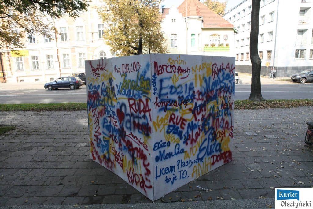 Kolorowa malowana na murze graffiti, pełna imion i dat, na tle miejskiego chodnika i drzew.