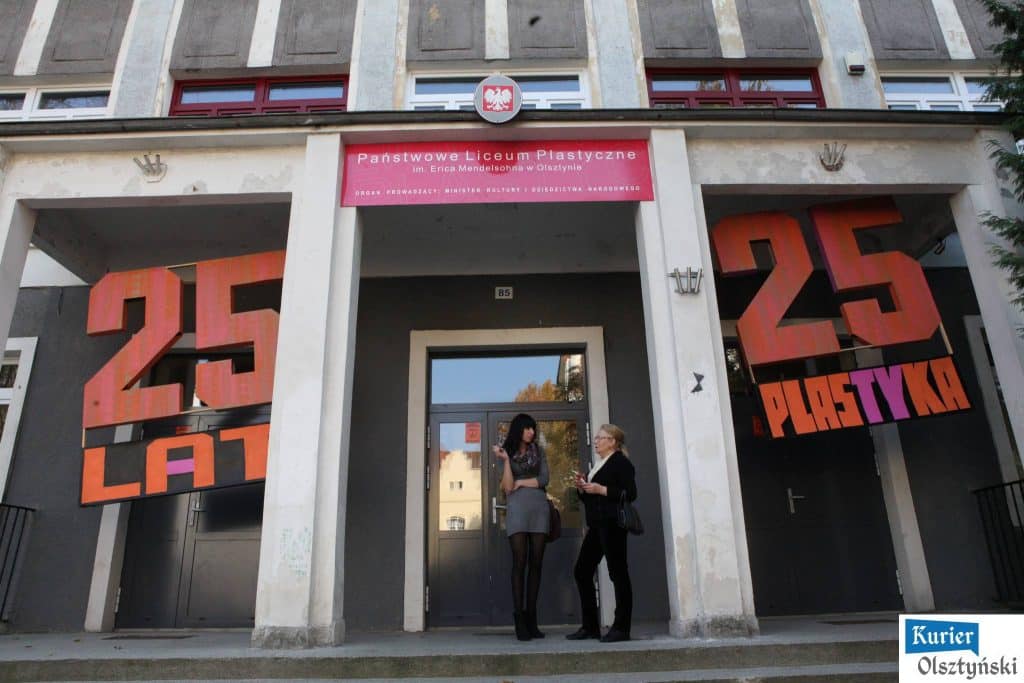 Dwie kobiet rozmawiają przed wejściem do budynku szkoły, na zewnątrz widoczne napisy i emblematy szkoły.