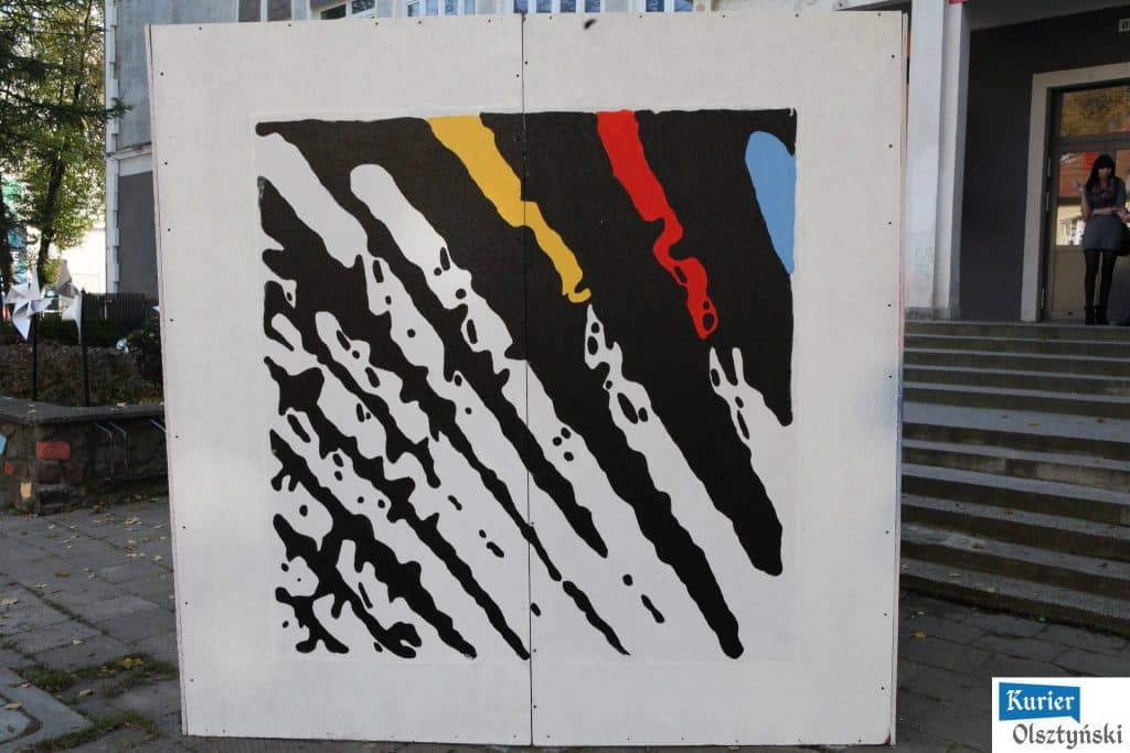 Abstrakcyjny mural na białej ścianie, w którym dominuje czarny kolor z akcentami żółtego, czerwonego i niebieskiego.