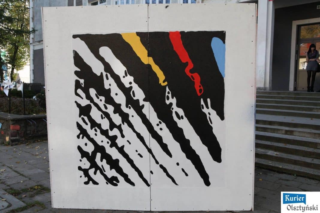 Abstrakcyjny mural na białej ścianie, w którym dominuje czarny kolor z akcentami żółtego, czerwonego i niebieskiego.