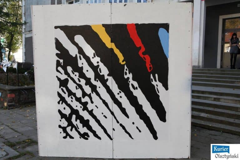 Abstrakcyjny mural na białej ścianie, w którym dominuje czarny kolor z akcentami żółtego, czerwonego i niebieskiego.