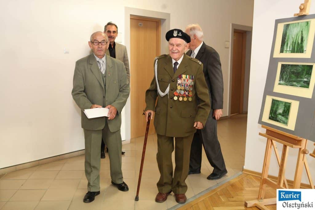 Grupa osób na otwarciu wystawy, w tym starszy mężczyzna w mundurze wojskowym z medalami, w towarzystwie innych mężczyzn w formalnych ubraniach.