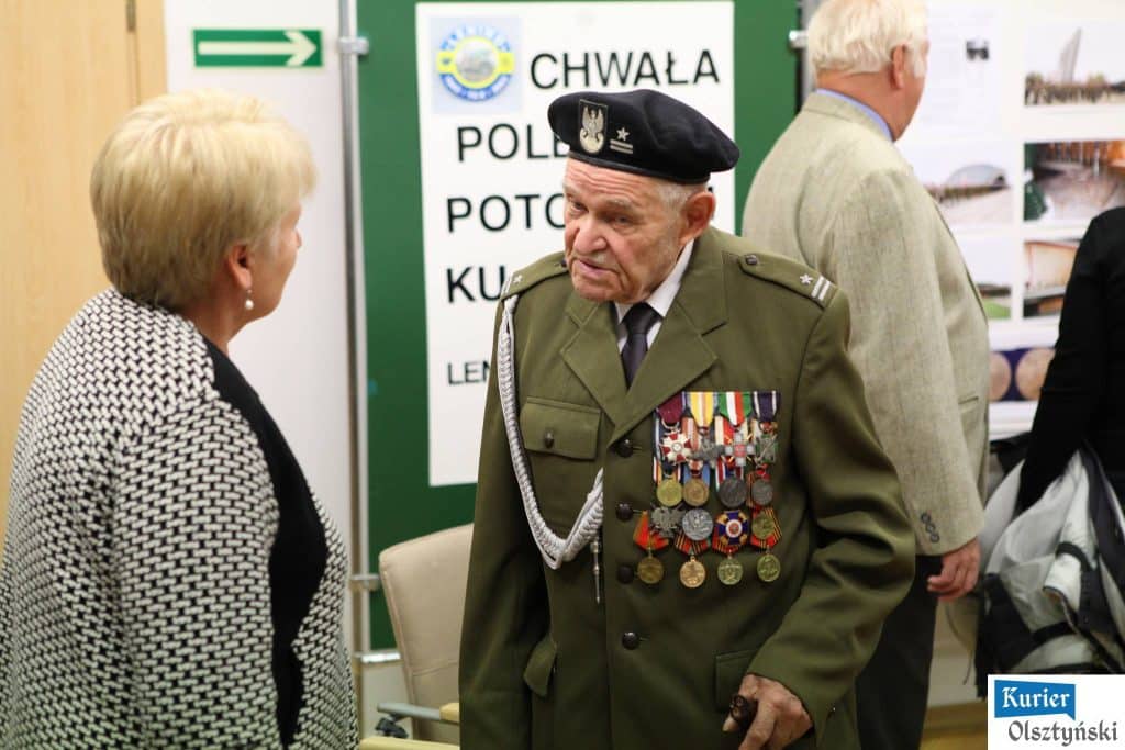 starszy mężczyzna w wojskowej uniformie z medalami, rozmawia z kobietą podczas wystawy w urzędzie.