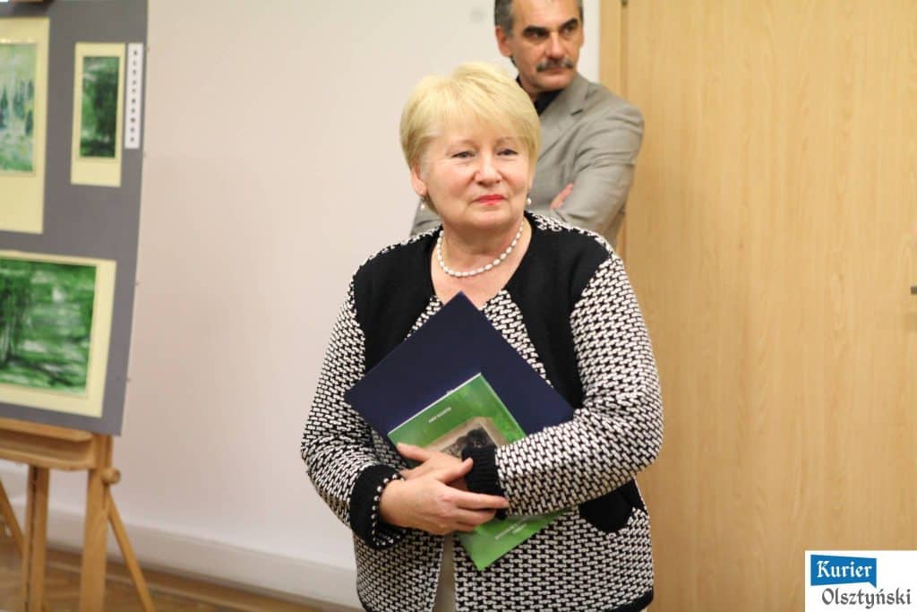 Kobieta trzymająca folder na wystawie zdjęć, w tle osoba stojąca przy ścianie, na obrazach widoczne pejzaże.