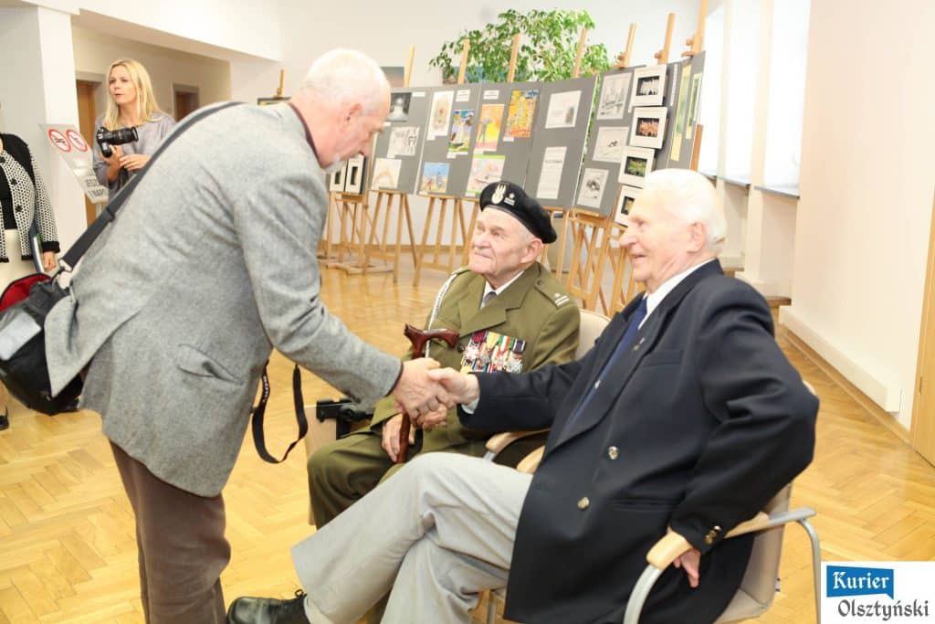 Dwie starsze osoby w mundurach wojskowych i cywilnych podczas spotkania na wystawie, rozmowa i uśmiechy.