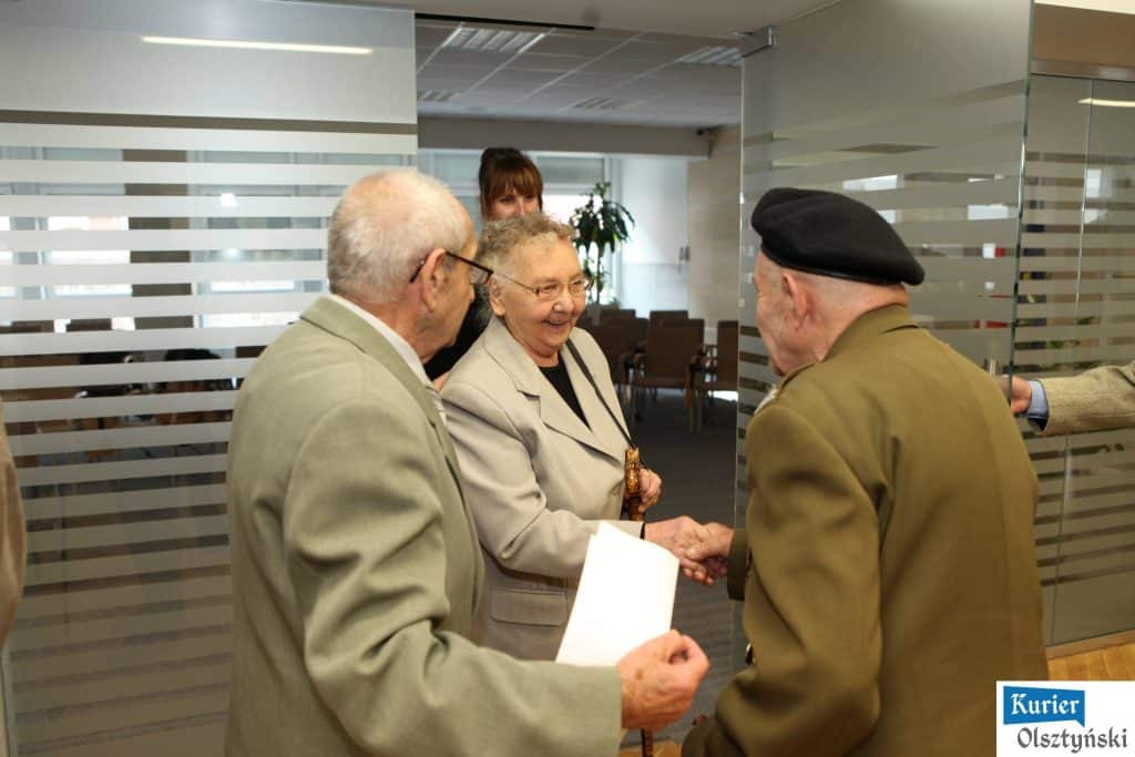 Grupa seniorów i seniorek rozmawia podczas otwarcia wystawy w urzędzie, w tle rośliny i sala konferencyjna.