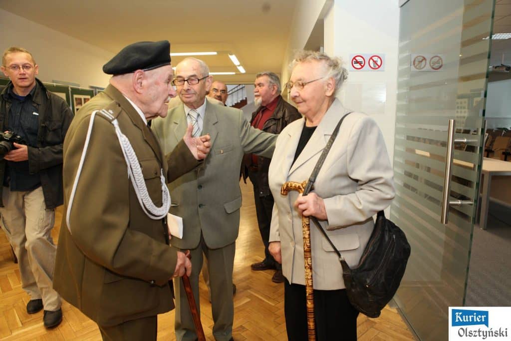 Grupa osób rozmawia podczas otwarcia wystawy w Urzędzie Wojewódzkim, wśród nich staruszek w mundurze wojskowym i starsza kobieta z laską.
