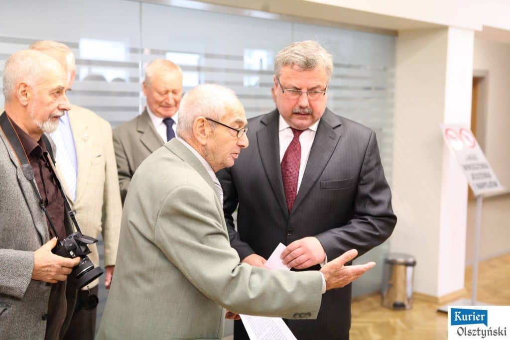 Grupa starszych mężczyzn na wystawie zdjęć w urzędzie wojewódzkim, jedna osoba pokazuje zdjęcie innym.