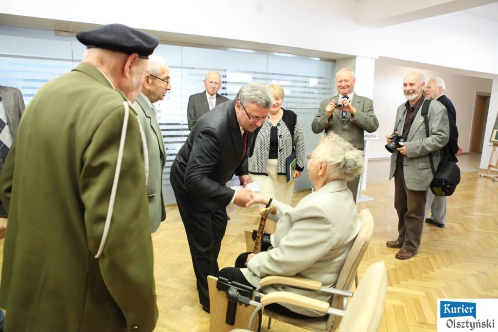 Grupa seniorów i działaczy w muzeum, jedna osoba siedzi z laską, inni jej składają życzenia.