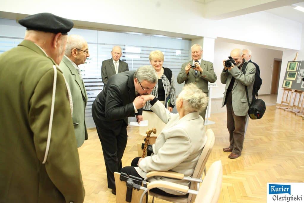 Urząd Wojewódzki, grupa ludzi podczas otwarcia wystawy, jedna osoba składa pocałunek na ręce starszej kobiety siedzącej na krześle, w tle fotografia i uśmiechnięte osoby.