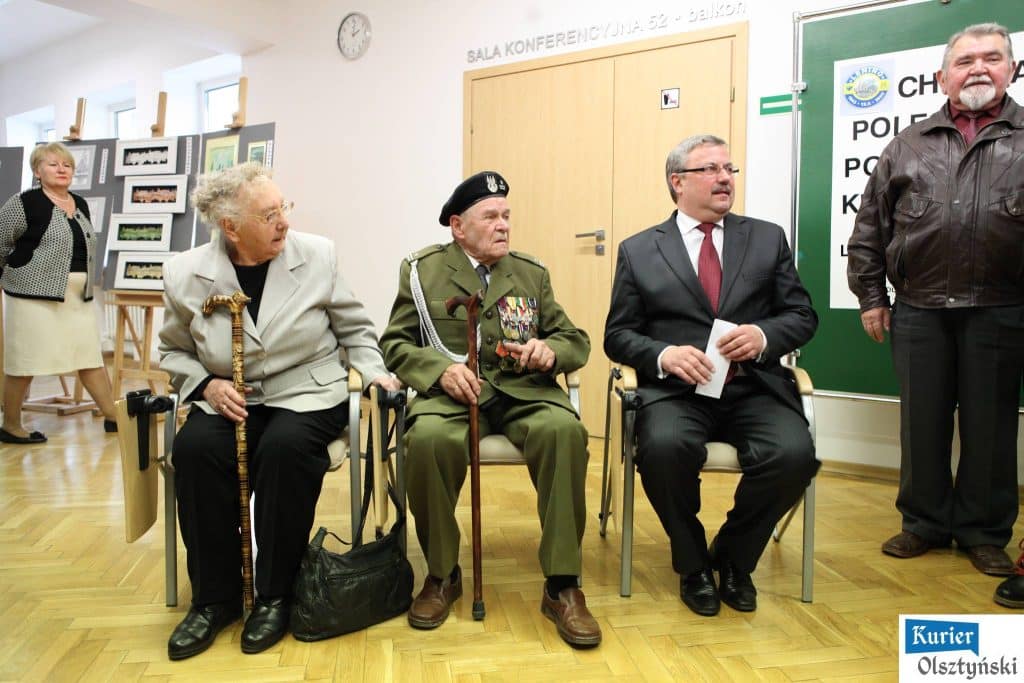 Grupa ludzi na wystawie poświęconej Bitwie pod Lenino, wśród nich starszy mężczyzna w mundurze wojskowym z medalami, kobiety i mężczyźni siedzą i stoją w pomieszczeniu.
