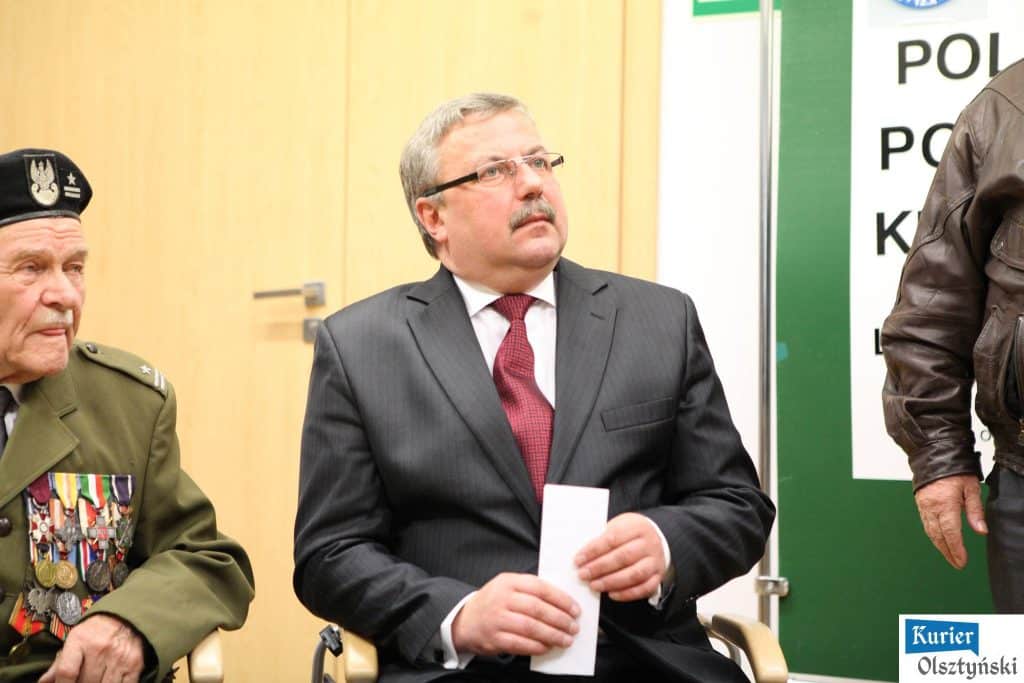Mężczyzna w garniturze i czerwonym krawacie, siedzi podczas wydarzenia, obok starszy mężczyzna w wojskowym mundurze z wieloma medalami.