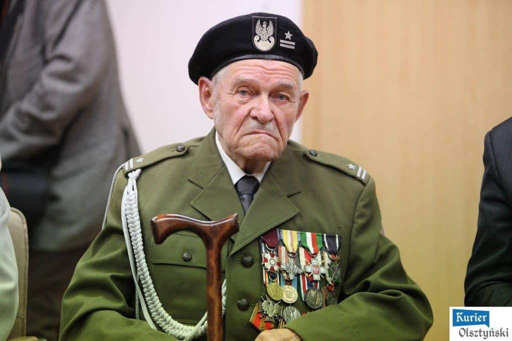 Starszy mężczyzna w mundurze wojskowym z medalami, na czapce ma odznakę wojskową, siedzi z poważnym wyrazem twarzy.