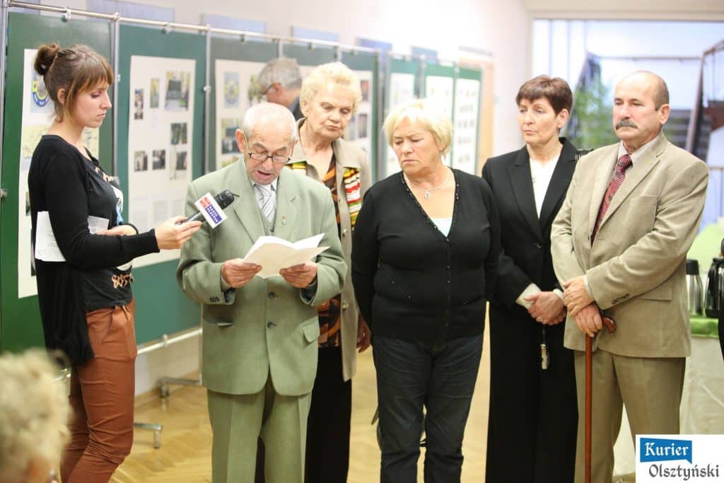 Grupa osób ogląda wystawę zdjęć w urzędzie wojewódzkim, w tym staruszek z okularami czytający z książki.