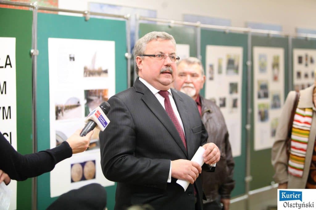 Na zdjęciu mężczyzna w garniturze i czerwonym krawacie podczas otwarcia wystawy w urzędzie wojewódzkim, za nim stoją ludzie przy tablicach z fotografiami.