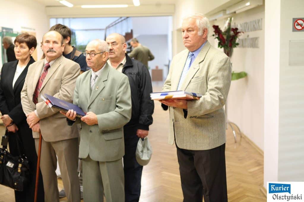 Grupa ludzi stojąca na wystawie w Urzędzie Wojewódzkim, z czego niektórzy trzymają albumy lub foldery, w tle rośliny i tablice.