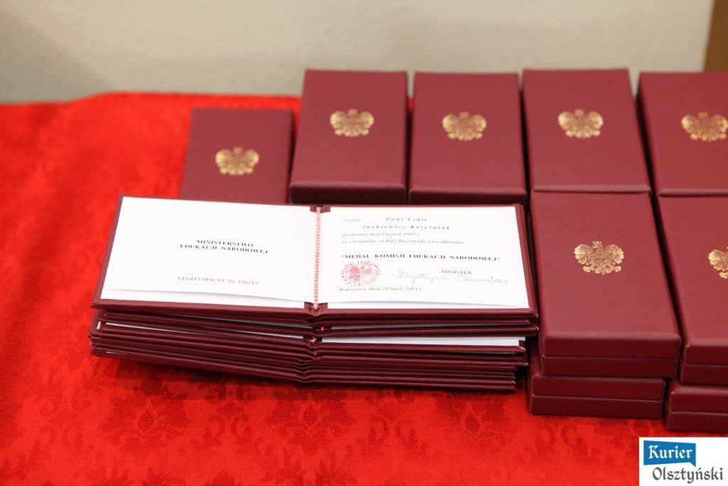 Na stole leżą odznaczenia i medaliki związane z Dniem Edukacji Narodowej, na tle czerwonej tacy z czterema prezentami.