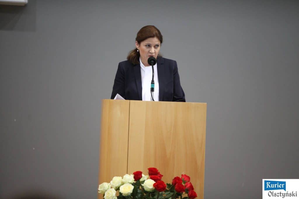 Kobieta przemawia z pulpitu podczas oficjalnych obchodów Dnia Edukacji Narodowej, za nią bukiet czerwonych i białych róż.