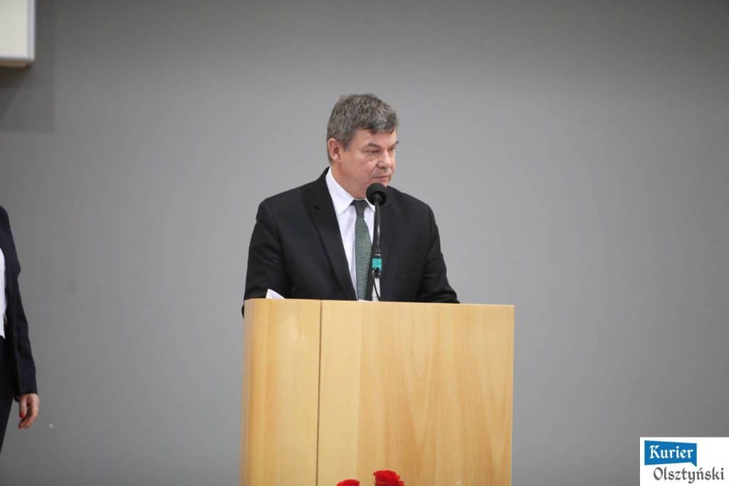 Mężczyzna w czarnym garniturze stoi za pulpitem podczas oficjalnych obchodów Dnia Edukacji Narodowej, na tle szarej ściany.