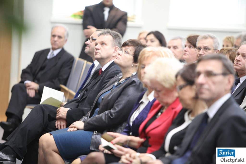 Uczestnicy uroczystości Dnia Edukacji Narodowej siedzą na sali, niektórzy czytają lub słuchają uważnie.