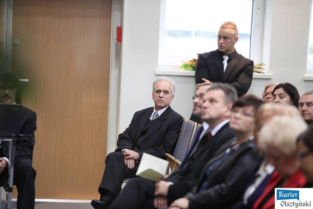Uczestnicy uroczystości Dnia Edukacji Narodowej w urzędzie wojewódzkim, wśród nich mężczyzna stojący z założonymi rękami, słuchają prelekcji.