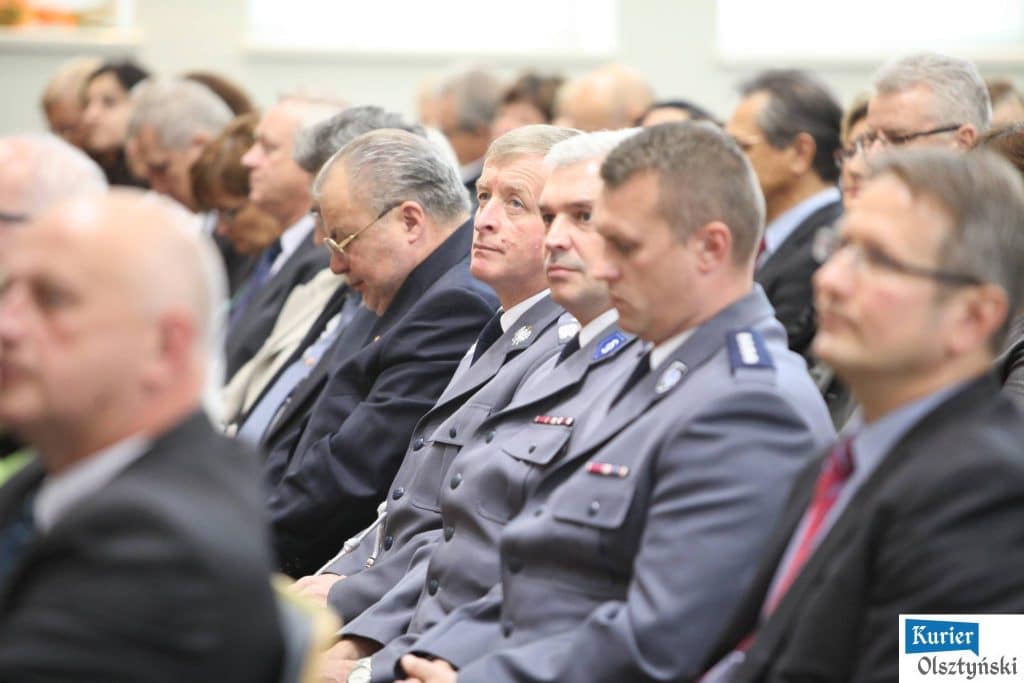 Uczestnicy oficjalnych obchodów Dnia Edukacji Narodowej w urzędzie, w tym osoby w mundurach i garniturach, siedzą na sali.
