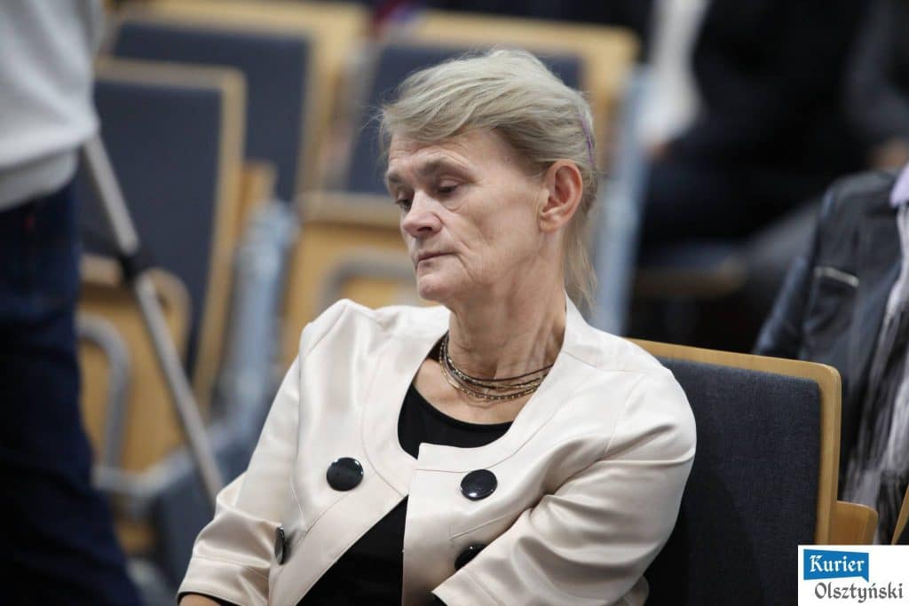 Starsza kobieta siedzi wśród innych uczestników na uroczystości Dnia Edukacji Narodowej, wygląda na zamyśloną.