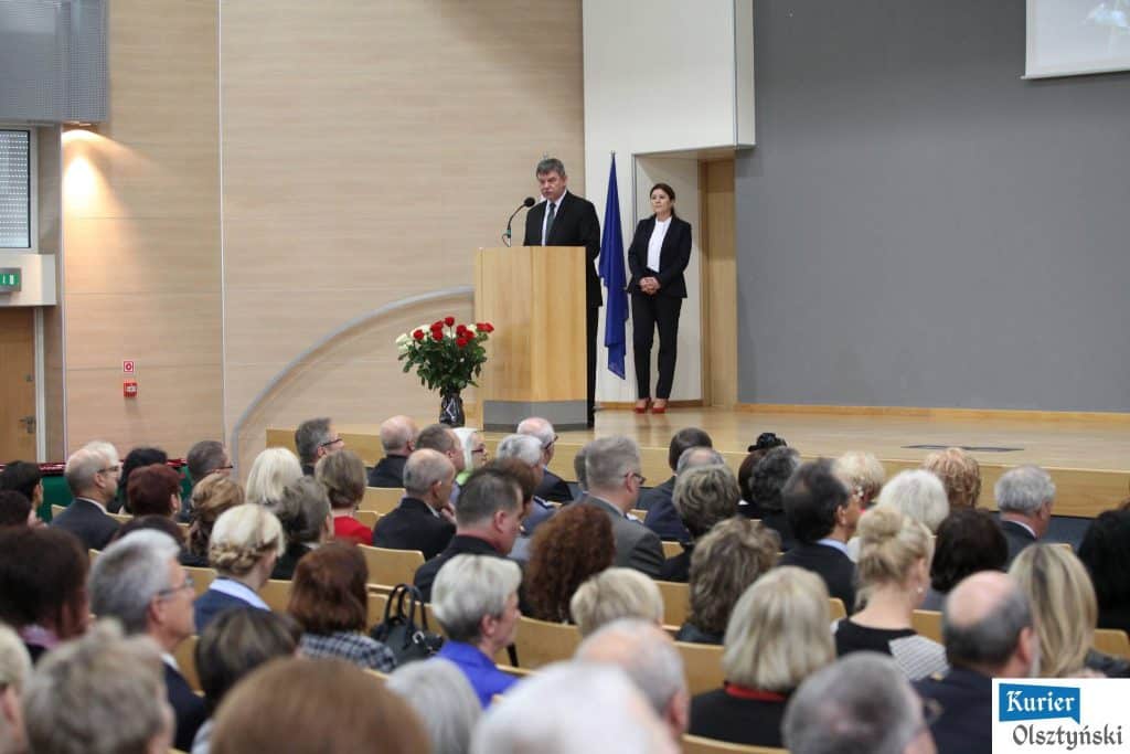 W uroczystości Dnia Edukacji Narodowej w Urzędzie Wojewódzkim przemawia mężczyzna przy mównicy, obok stoi kobieta. Na scenie stoją flagi i kwiaty.