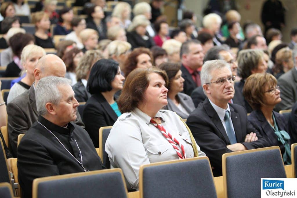Uczestnicy uroczystości Dnia Edukacji Narodowej siedzą na sali, słuchając wystąpień. W centrum kobieta w uniformie z przypinkami.