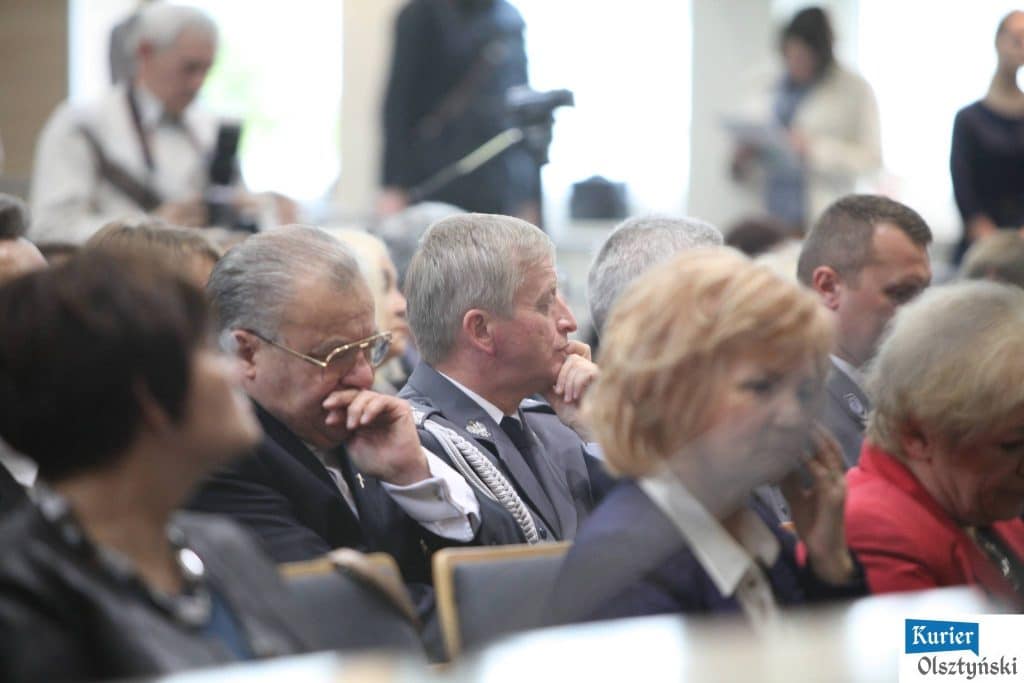Uczestnicy uroczystości Dnia Edukacji Narodowej siedzą na sali, słuchając prezentacji, na pierwszym planie widoczne osoby w sutannach i mundurach.